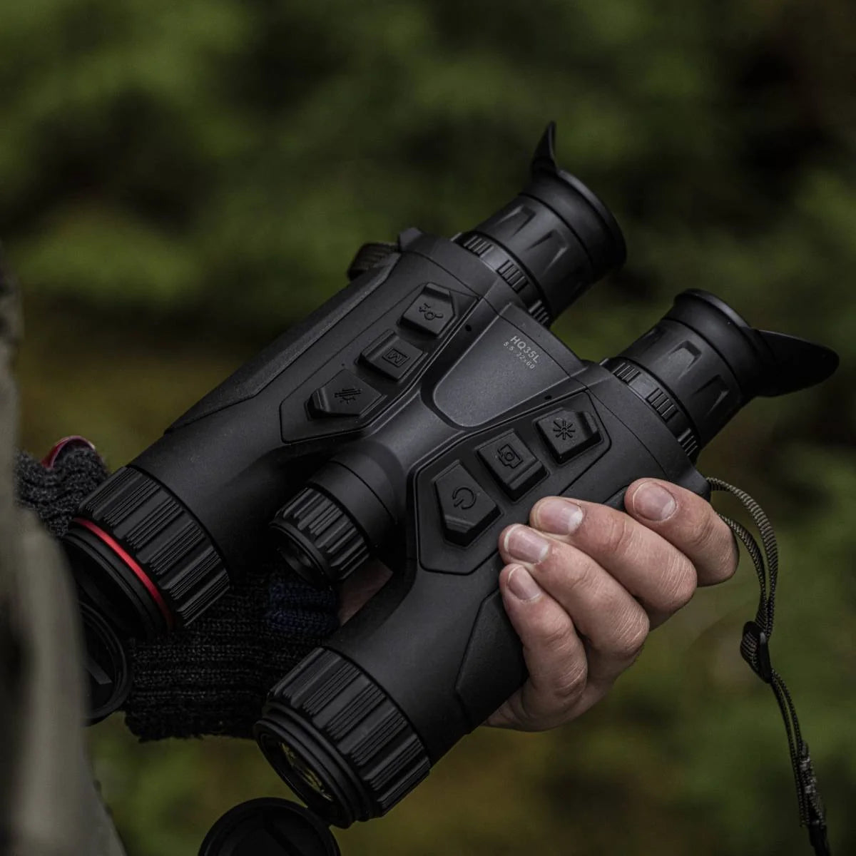 Una mano sostiene un binocular multiespectral Hikmicro HQ35L 5.5-22x60 en terreno, con vegetación desenfocada al fondo.