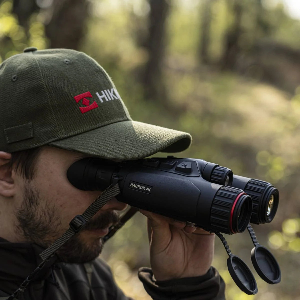 Una persona con gorra observa a través de un binocular multiespectral Hikmicro HQ35L 5.5-22x60, de perfil, en un bosque.